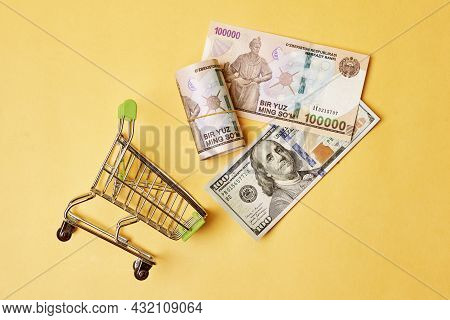Buying Us Dollars In Uzbekistan. Us Dollars Roll In Shopping Trolley And Pile Of Uzbek Sums. Uzbek M