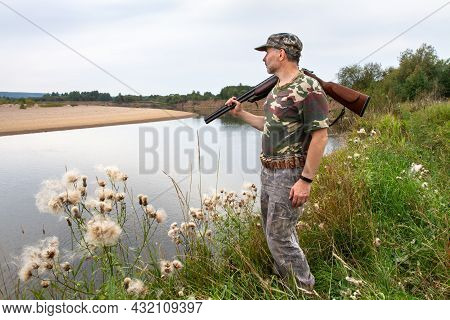 The Hunter Stands On The Bank And Looks At The River. He Carries An Unloaded Shotgun On His Shoulder