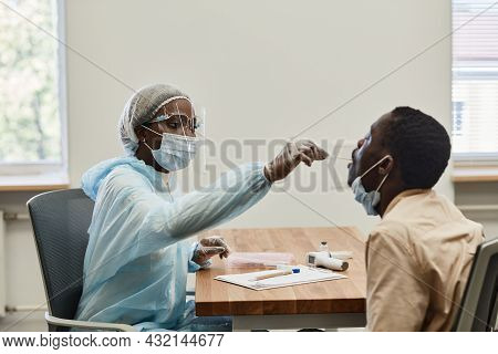 Black Medical Nurse In Protective Suit And Face Shield Asking Patient To Open Mouth So She Could Get