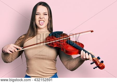 Young hispanic girl playing violin sticking tongue out happy with funny expression. 