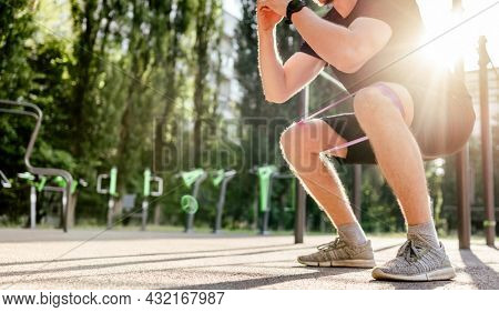 Strong sportive man guy doing squats with elastic rubber band at the stadium outdoors. Adult male person during workout for legs with additional sport equipmant. Sportsman exercising outside