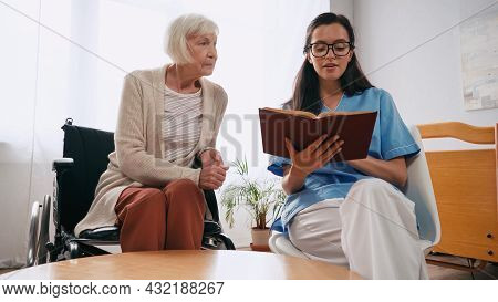 Brunette Geriatrician Nurse Reading Book To Senior Woman In Wheelchair