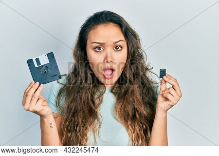Young hispanic girl holding floppy disk and sdxc card in shock face, looking skeptical and sarcastic, surprised with open mouth 