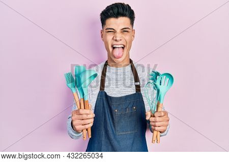 Young hispanic man wearing professional baker apron holding cooking tools sticking tongue out happy with funny expression. 