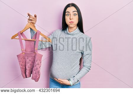 Beautiful woman with blue eyes expecting a baby holding baby clothes puffing cheeks with funny face. mouth inflated with air, catching air. 