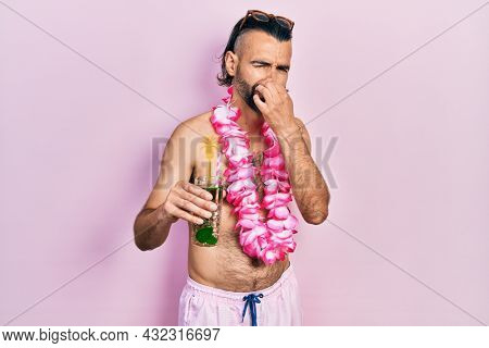 Young hispanic man wearing swimsuit and hawaiian lei drinking tropical cocktail smelling something stinky and disgusting, intolerable smell, holding breath with fingers on nose. bad smell 