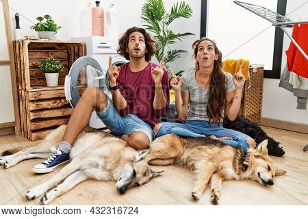 Young hispanic couple doing laundry with dogs amazed and surprised looking up and pointing with fingers and raised arms. 