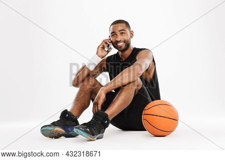 Happy african young basketball player sitting talking on mobile phone isolated on a white background
