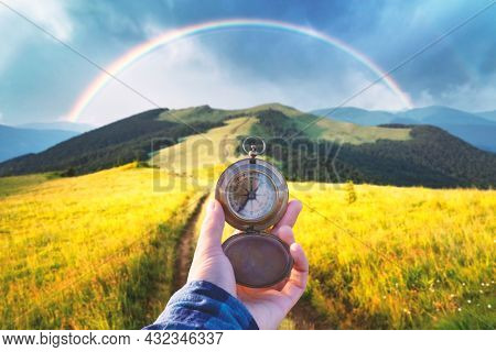 Man with old metal compass in hand on mountains road. Travel concept. Lush green grassy meadows and beautyful rainbow on background. Landscape photography