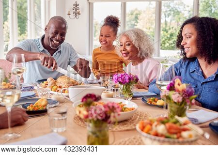Grandfather Carving As Multi Generation Family Sit Around Table At Home And Enjoy Eating Meal