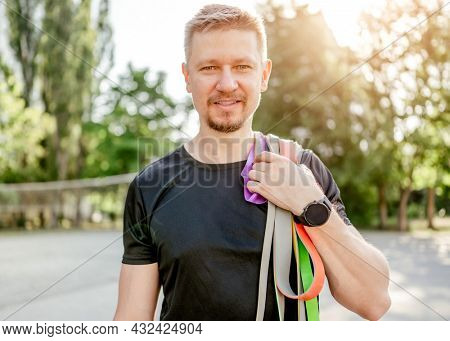 Sportive man guy holding in his hand set of colorful elastic rubber bands and smiling looking at the camera at the stadium outdoors. Athlete male person during workout with additional sport equipmant
