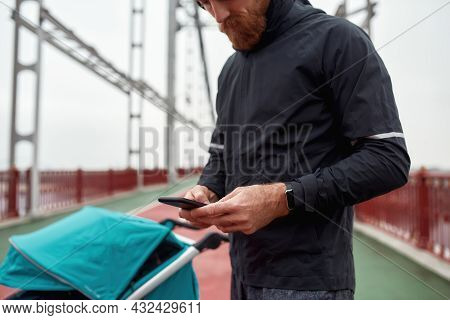 Cropped Shot Of Male Runner Dad Using Smartphone While Preparing For A Jog In The City With Stroller