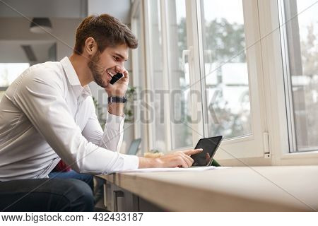 Young smiling european businessman talking on mobile phone and using digital tablet. Concept of modern successful man. Bearded stylish guy wearing formal clothes. Office interior. Daytime