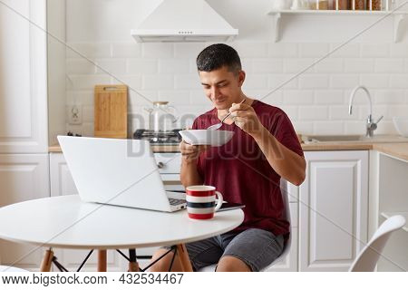 Hungry Man Freelancer Sitting In Kitchen At Table In Front Of Open Laptop Computer And Eating Soup, 