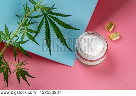 Hemp Face Cream In A Glass Jar, Two Cbd Oil Capcules And And Green Cannabis Sativa Leaves Against Pi