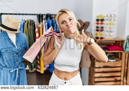 Young caucasian woman holding shopping bags at retail shop cutting throat with hand as knife, threaten aggression with furious violence 