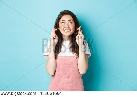 Portrait Of Hopeful Brunette Girl Cross Fingers For Good Luck, Making Wish, Looking Up With Begging 