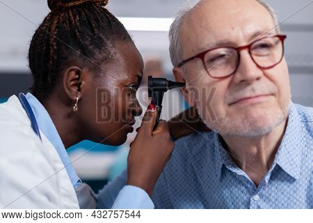 Close Up Of Black Doctor Using Otoscope For Ear Checkup Examination On Old Patient In Cabinet. Afric