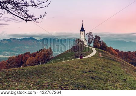 Beautiful Sunset Landscape Of Church Jamnik In Slovenia On Green Hill With The Trees And Pink Sky