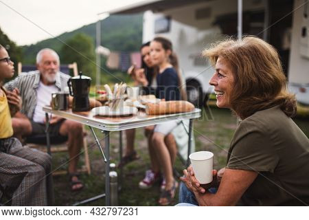 Multi-generation Family Sitting And Eating Outdoors By Car, Caravan Holiday Trip.