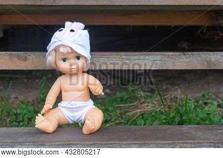 Cute Kewpie Doll In Hat And Shorts Sits On Rustic Wooden Bench In Country House Yard Space For Text.