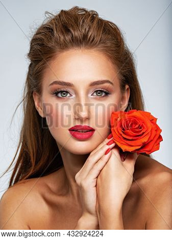Beautiful young woman with a red flower in hand near face.  Portrait of white girl with red rose in hands. Beauty face concept. Art portrait of an attractive model. Girl with bright fashion makeup