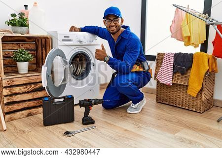 Young indian technician working on washing machine doing happy thumbs up gesture with hand. approving expression looking at the camera showing success. 