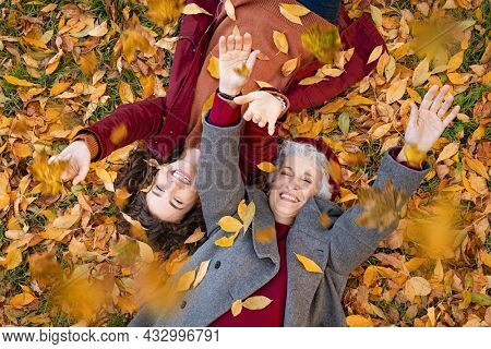 Senior grandmother and smiling granddaughter lying on yellow and red leaves at park, view from above. Top view of playful old woman with cheerful girl lying on autumn park leaves, looking at camera.