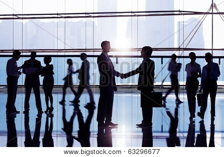 Two businessmen shaking hand in the office building admist other business people.