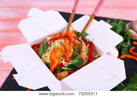 Chinese noodles and sticks in takeaway box on black mat on pink background