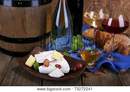 Supper consisting of Camembert and Brie cheese, honey, wine and grapes on napkin in basket and wine barrel on wooden table on brown background