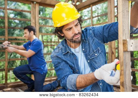 Male carpenter measuring wooden plank with tape while colleague working in background at construction site