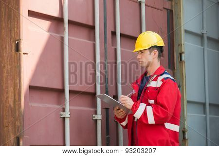 Male worker inspecting cargo container while writing on clipboard in shipping yard