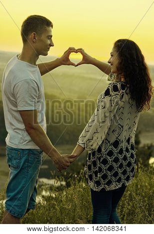 romantic couple at sunset make a heart shape from hands, beautiful landscape and evening sky, love tenderness concept, young adult people, yellow toned