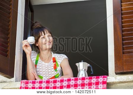 Portrait of young woman is enjoying a cup of freshly brewed coffee with Italian moka pot sitting near open window with traditional European wood brown shutters.