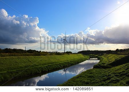 A wind turbine in a peaceful landscape with green grass blue skye and a river
