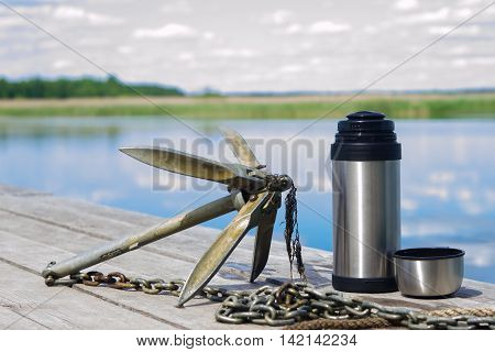 Anchor with a thermos of tea on a background of nature are at the pier