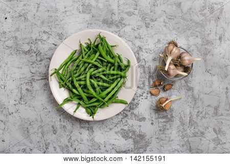 French bean in white plate on the rough light gray surface with garlic top view. Ingredients for making helpful vegetarian food