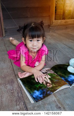Closeup asian girl smiling and reading picture album lying on wooden floor with bright sunlight outdoor at home. Children read and study education concept.
