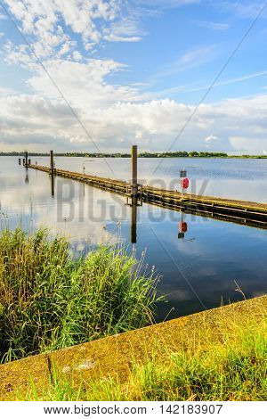 Long wooden jetty in a Dutch lake with a mirror smooth water surface and diagonally into the image. It is early in the morning on a beautiful windless summer day.