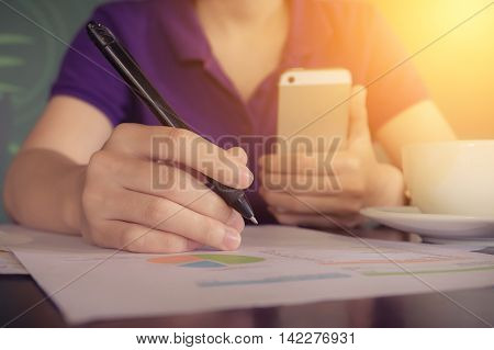 close up hand of business women holding pen pointing on paper garph chart and using smart phone on wooden desk indoor office with morning light. vintage filter effect.