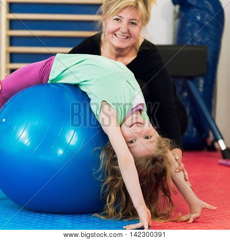 Physical therapist working with little girl in school gymnasium exercising with fitness ball