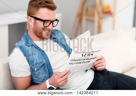 In pace with the world. Cheerful content handsome smiling man sitting on the couch and resting while resting newspaper