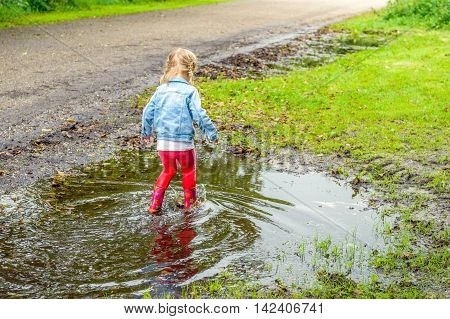 Little toddler girl with blonde pigtails en red boots walking through the water in the puddle. By playing in this way she discovers the world gradually.