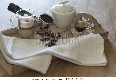 Wooden tray with glass of latte macchiato, espresso, grouphead, coffee beans and brown sugar