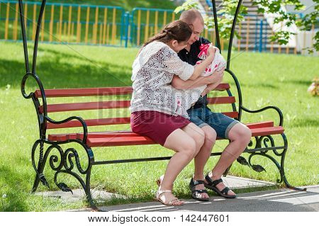 Young Family Sitting On A Park Bench. Father Holding Newborn Daughter In Her Arms.
