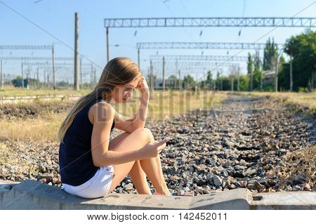 Girl teenager in top and shorts using cellphone while sitting on concrete of unfinished rail track in the countryside