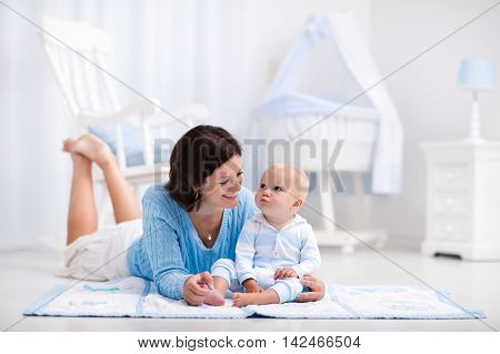 Happy young mother and adorable baby boy playing on a blue floor mat in a white sunny nursery with rocking chair and bassinet. Bedroom interior with infant crib. Mom and child on playmat at kids bed.
