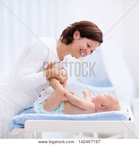 Young mother taking care of little boy in white sunny nursery with changing table baby crib and rocking chair. Diaper change and clothing. Mom and son in beautiful bedroom at home.