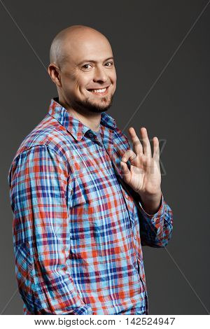 Portrait of confident handsome cheerful middle-aged man in plaid shirt smiling at camera showing ok gesture over grey background. Copy space.